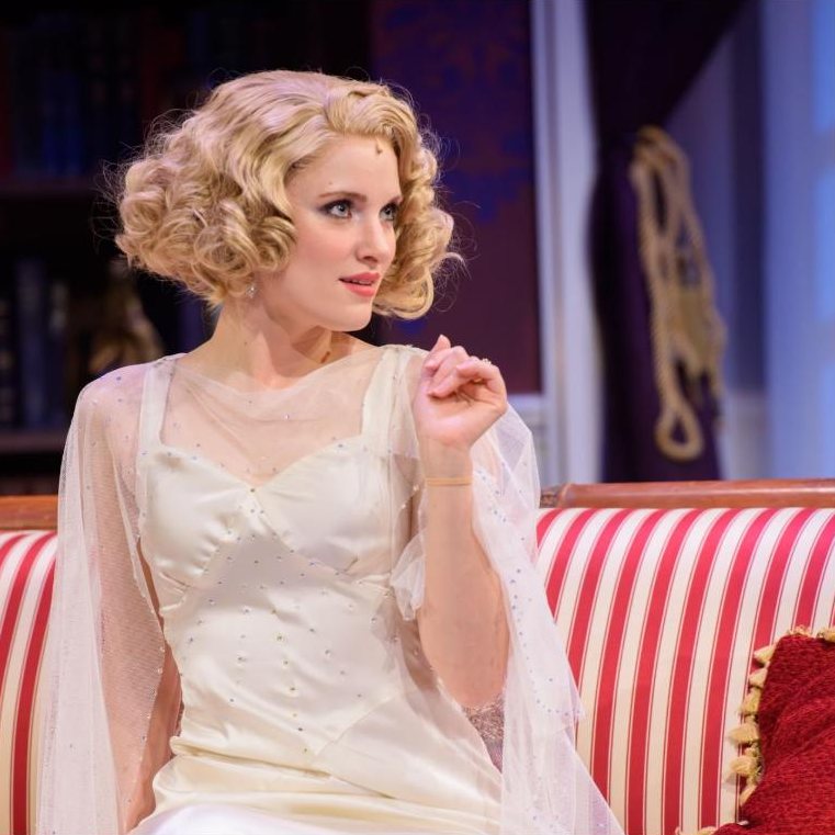 Paige as Elvira in a white beaded gown and finger-waved blonde wig, seated on a striped sofa