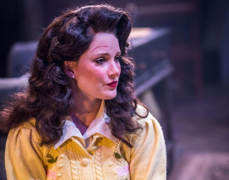 Paige as Susan Hendrix with dark 1940s curls and a yellow cardigan, looking off stage